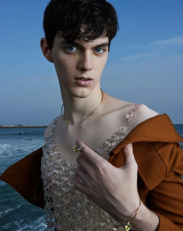 Adult male model poses outdoors wearing a sheer beaded top, gold necklace, ring, and a brown jacket with an ocean backdrop.