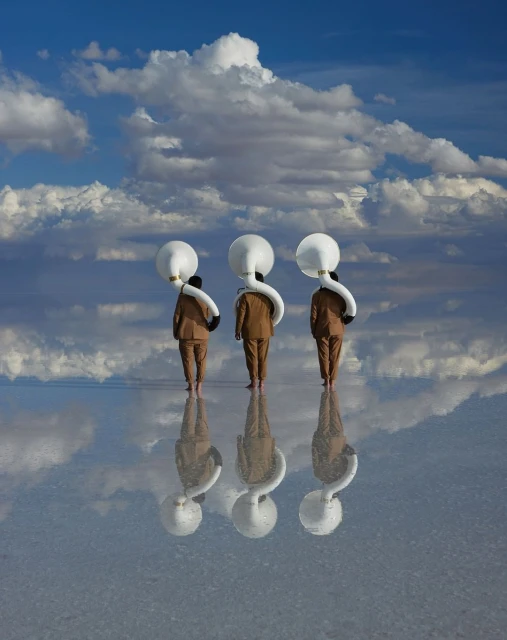 Three figures in tan suits stand on a reflective salt flat under a blue sky with white clouds, each wearing a white, bulbous headpiece.