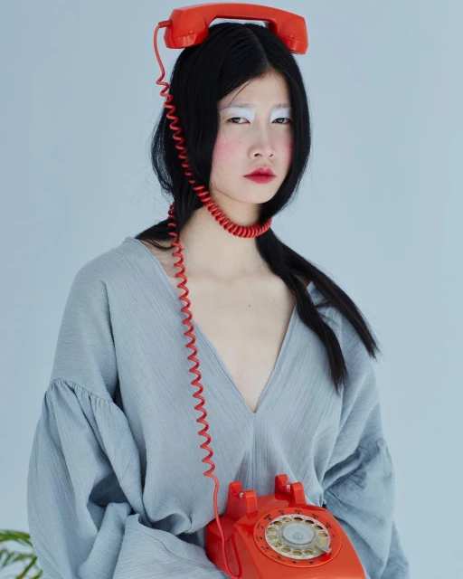 Artistic portrait of a young Asian woman with black hair, styled with vintage red telephones on her head and in her lap.
