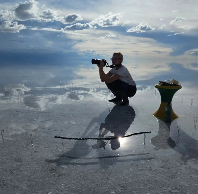 A woman with glasses and a camera squats in a vast, shallow body of water reflecting a cloudy sky. A small table is to the right.