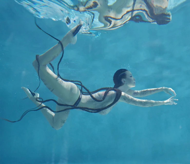 Underwater shot of an adult woman swimming in a pool with long black braids, wearing a light-colored swimsuit, photographed at eye-level.