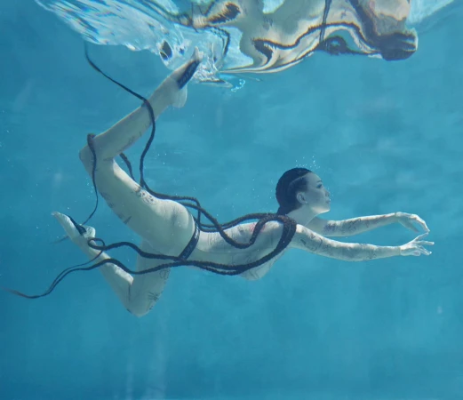 Underwater shot of an adult woman swimming in a pool with long black braids, wearing a light-colored swimsuit, photographed at eye-level.