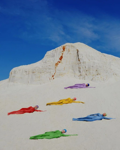 Five people in colorful bodysuits lying on a bright white sand dune under a clear blue sky, creating a visually striking scene.