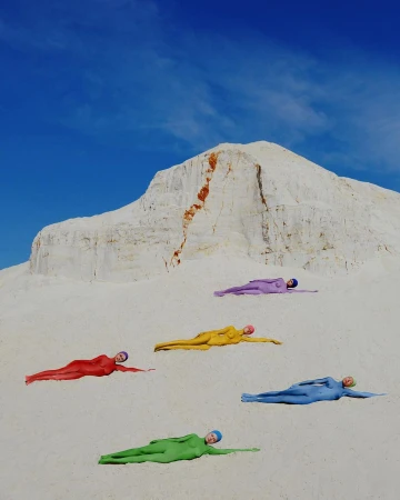 Five people in colorful bodysuits lying on a bright white sand dune under a clear blue sky, creating a visually striking scene.