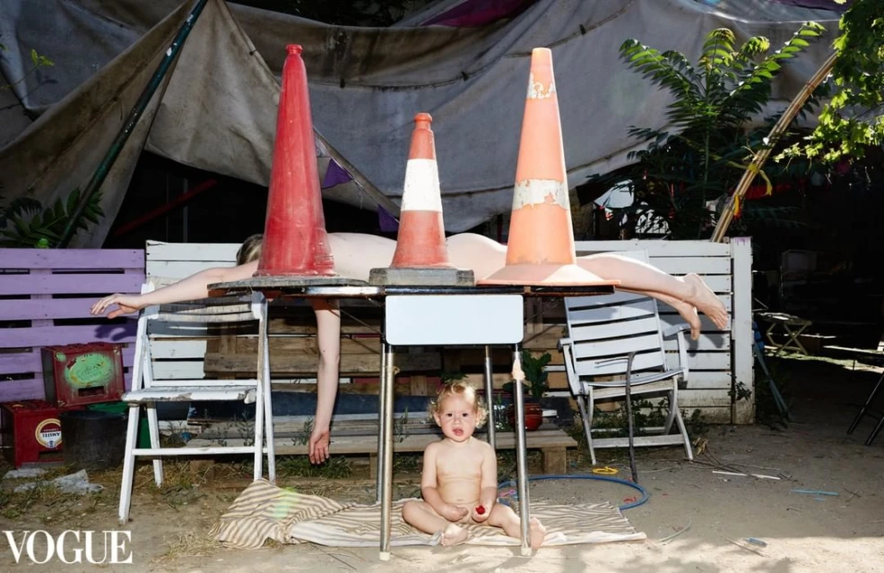 Conceptual outdoor scene features a child, a reclining nude adult, and traffic cones arranged on tables, creating a surreal composition.