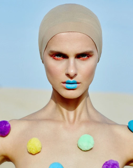 Close-up of a woman with a head covering, bright blue lipstick, and colorful pom poms attached to her shoulders against a desert background.