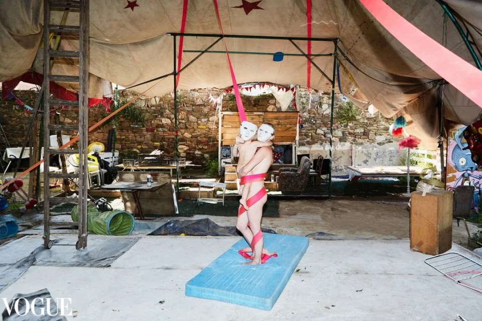 Artistic shot inside a tent with two figures wearing white masks and wrapped in a pink ribbon, standing on a blue mattress pad.