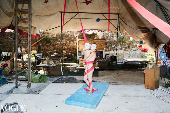 Artistic shot inside a tent with two figures wearing white masks and wrapped in a pink ribbon, standing on a blue mattress pad.