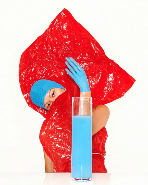 Eye-level, indoor studio shot with an Asian woman in a blue cap and gloves, partially concealed by a red plastic sheet and blue liquid in a glass.
