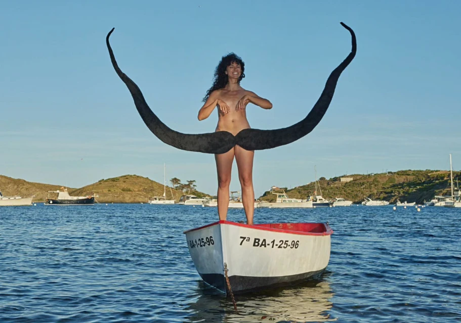 Nude woman stands in a rowboat holding giant mustache against a background of blue water, boats, and green hills under a blue sky.
