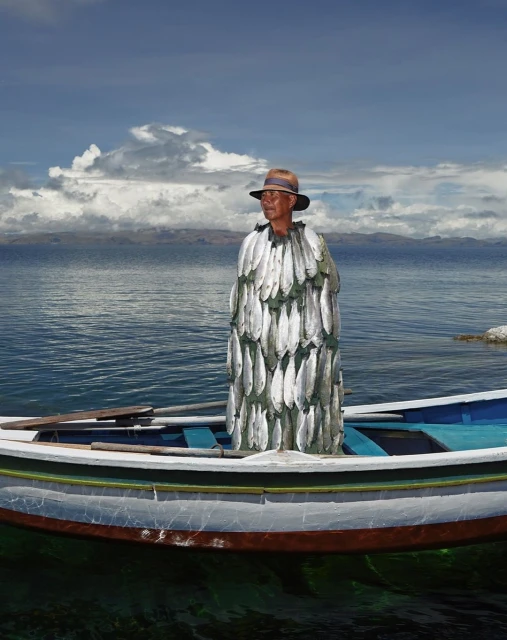 An adult man wearing a hat and cape made of fish sits inside of a blue and white boat on a lake with cloudy skies above.