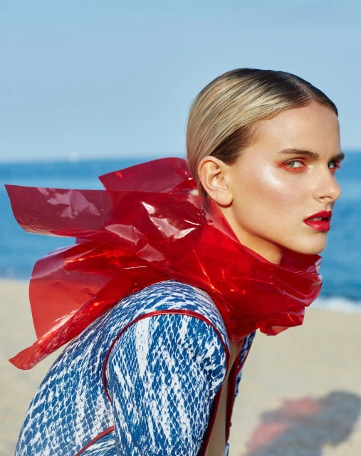 A woman with red eye makeup and lipstick poses outdoors wearing a blue and white patterned shirt with a transparent red plastic collar.