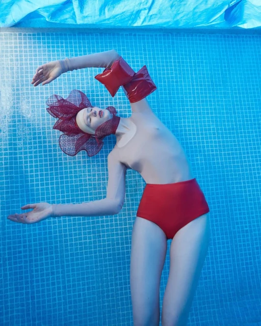 Woman floats in blue-tiled pool wearing red high-waisted bottoms and sculptural red mesh floral headpiece and matching wrist adornments.