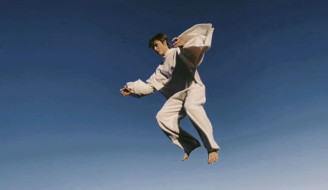 Man in a cream-colored outfit leaps mid-air against a vivid blue sky in a dynamic, low-angle shot capturing movement and fashion.