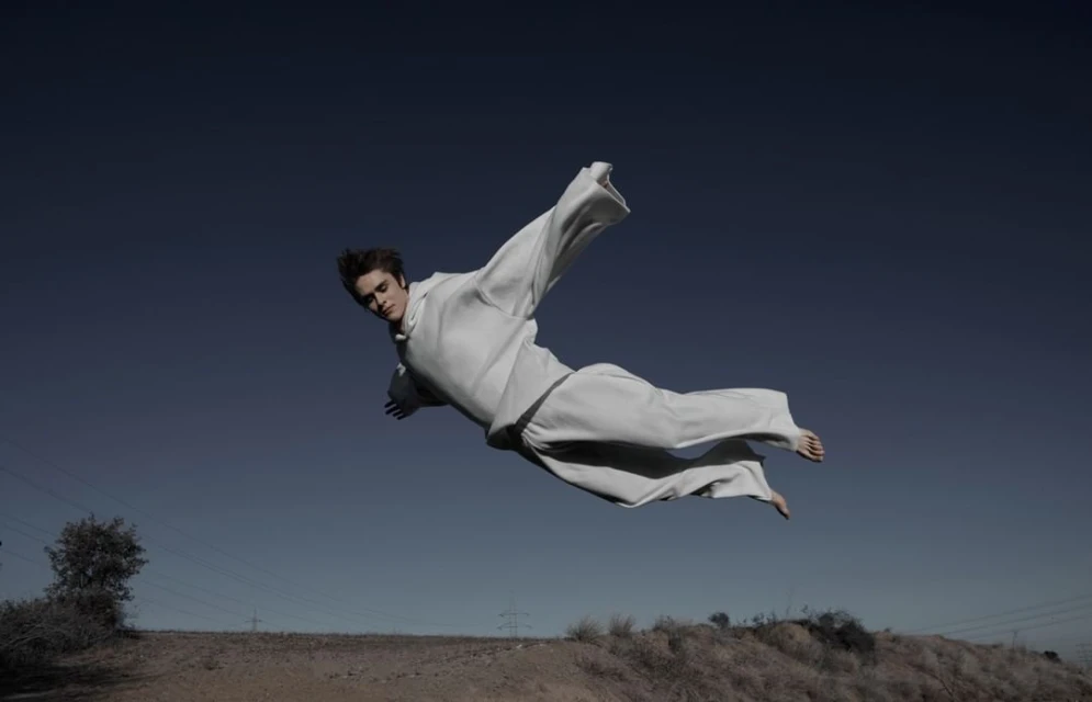 Man in white robe floating in the air against a blue sky, landscape in the background, studio photography with dramatic lighting.