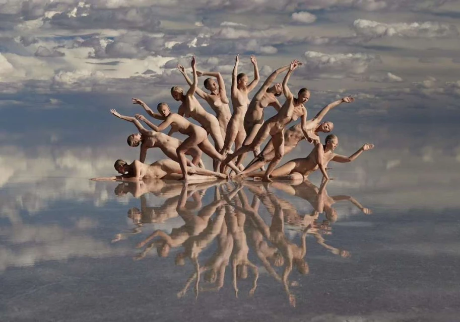 A group of nude female figures are arranged in a unique formation on a reflective surface, with a cloudy sky backdrop enhancing the artistic scene.