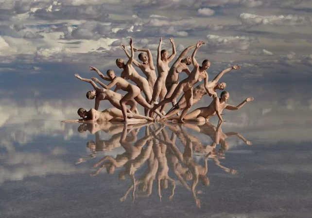 A group of nude female figures are arranged in a unique formation on a reflective surface, with a cloudy sky backdrop enhancing the artistic scene.