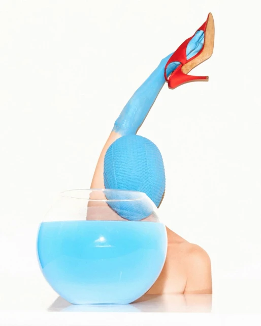 Abstract fashion image: arm painted blue holds blue ball above blue glass bowl with red high-heeled shoe balanced on painted fingers.