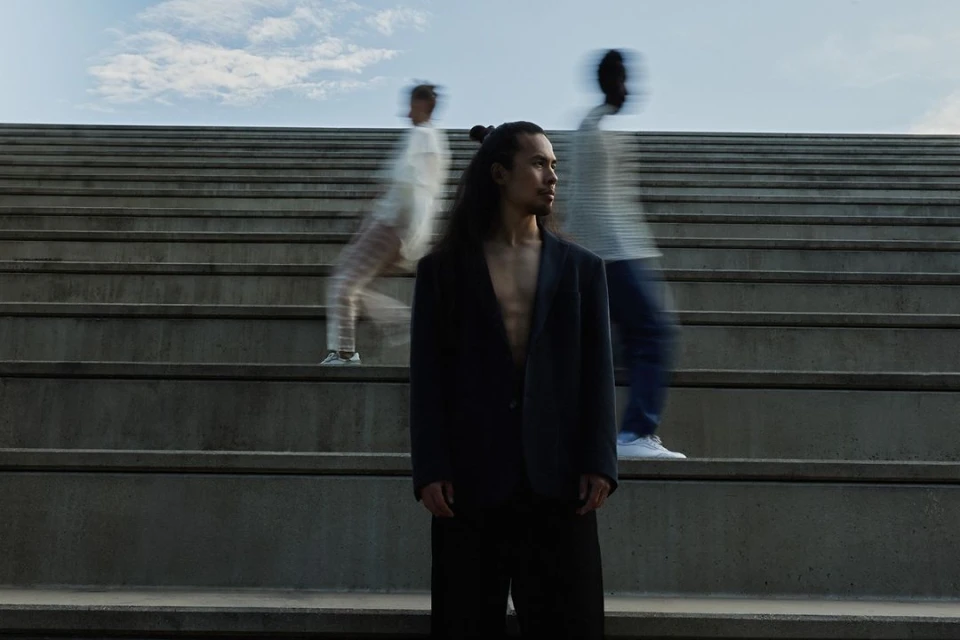 Man in a blazer stands on concrete stairs, flanked by blurred figures, with a cloudy sky in the background.