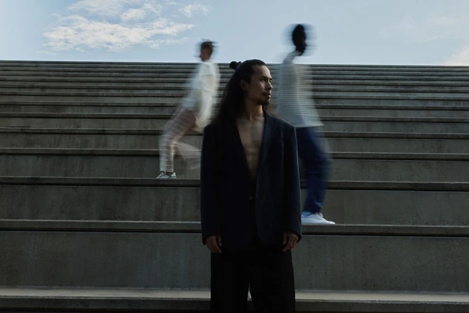 Man in a blazer stands on concrete stairs, flanked by blurred figures, with a cloudy sky in the background.