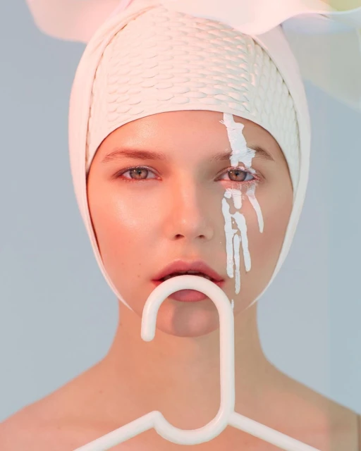 Close-up portrait of a woman with white paint dripping from her eye, wearing a white head covering and holding a white hanger.
