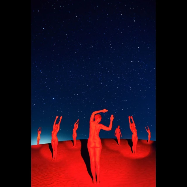 A group of nude, red-toned figures stand on a sandy surface under a dark blue, star-filled night sky in an artistic photograph.