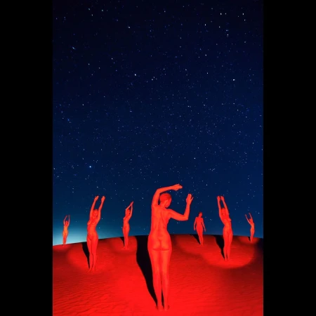 A group of nude, red-toned figures stand on a sandy surface under a dark blue, star-filled night sky in an artistic photograph.
