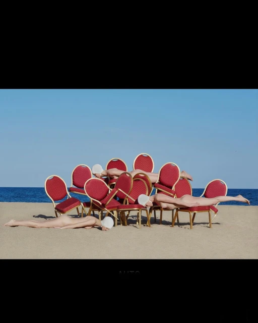 Eleven nude humans wearing white swim caps lie on or near red upholstered chairs on a sandy beach under a clear blue sky.