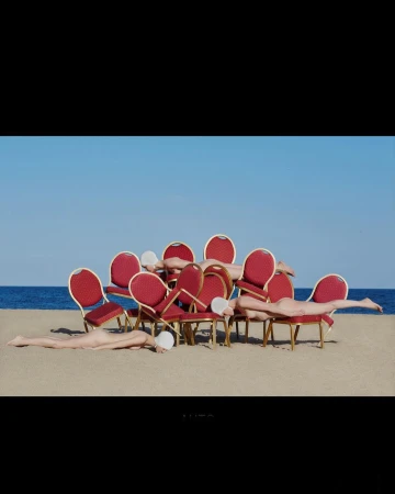 Eleven nude humans wearing white swim caps lie on or near red upholstered chairs on a sandy beach under a clear blue sky.