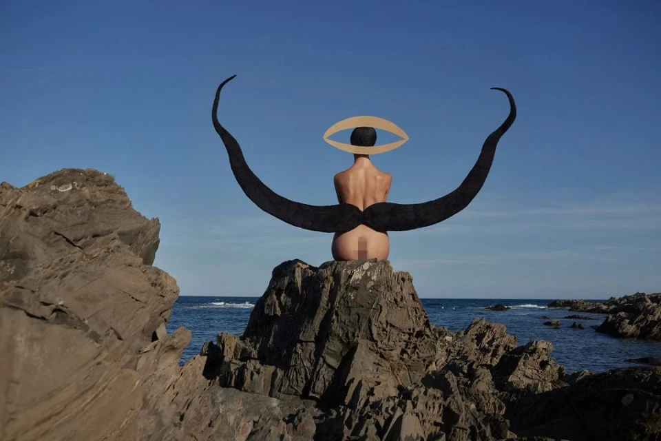 Artistic nude woman seated on rocks near ocean, featuring eye graphic and large moustache-shaped headpiece, against clear blue sky.