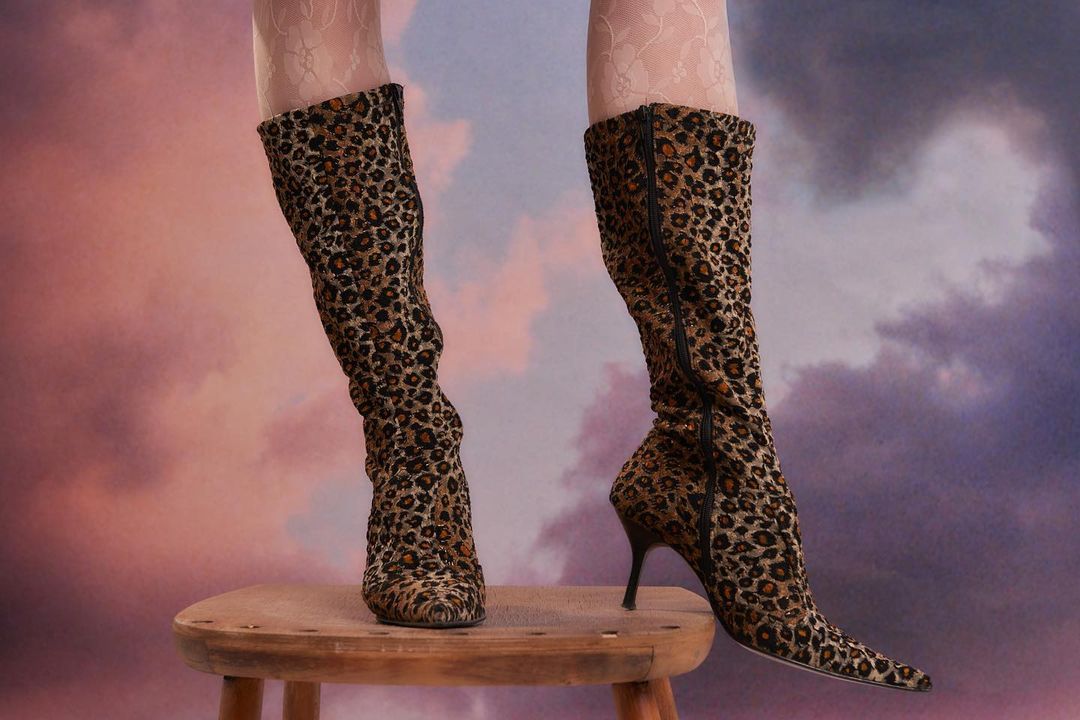 Leopard print knee-high boots are showcased on a wooden stool against a cloud-painted backdrop, accented by sheer floral lace stockings.