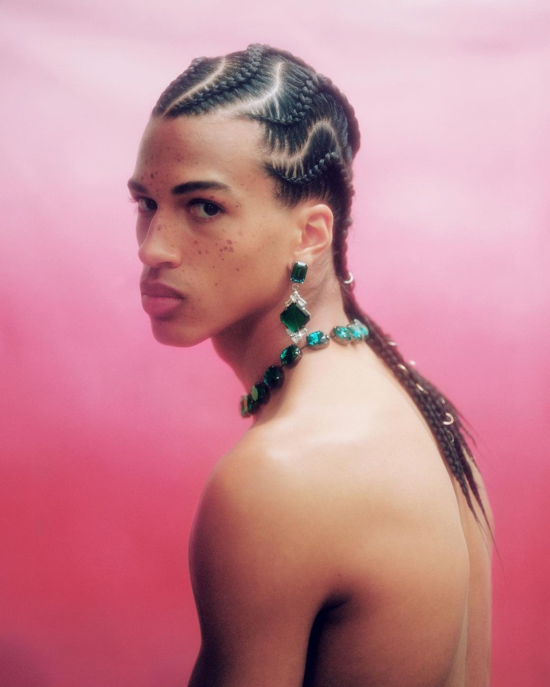Headshot of a Black male model with cornrow braids, wearing large emerald earrings and a matching necklace against a pink gradient background.