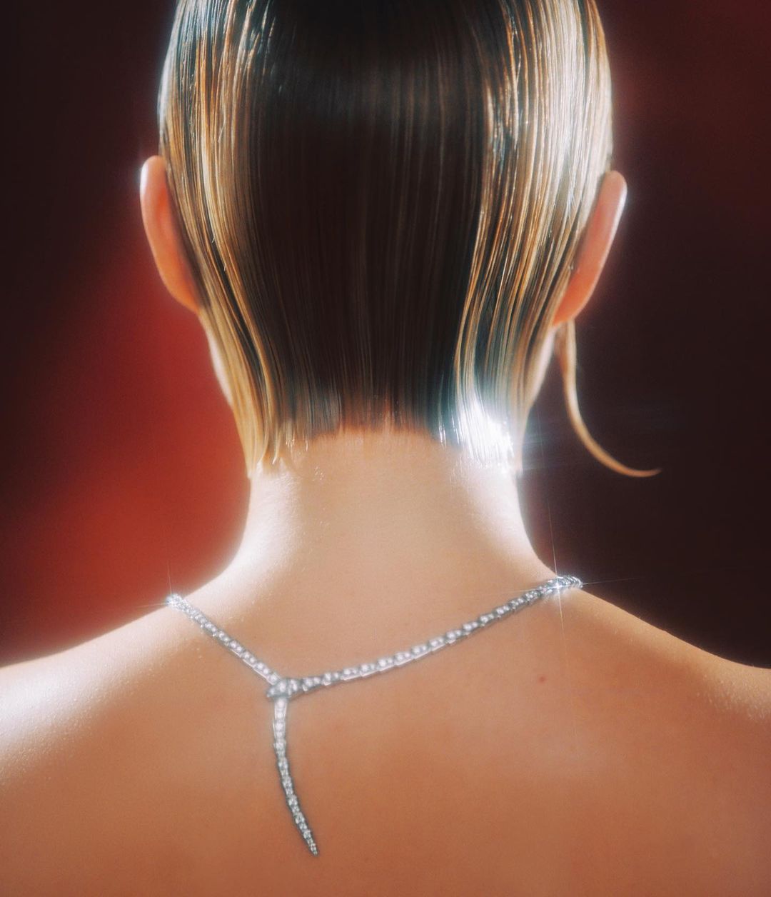 Woman with blonde hair and bare back wearing a diamond necklace in front of a red background in a medium shot with studio lighting.