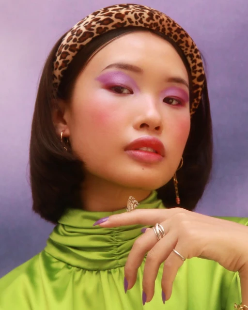 Asian woman in a green high-neck blouse with leopard-print headband and bright purple makeup posing indoors for a beauty portrait.
