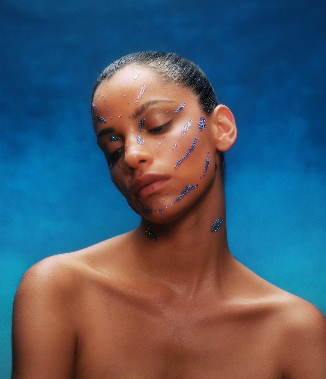 Portrait of a woman with glitter on her face against a blue background. Her eyes are closed and her face is serene in a studio setting.