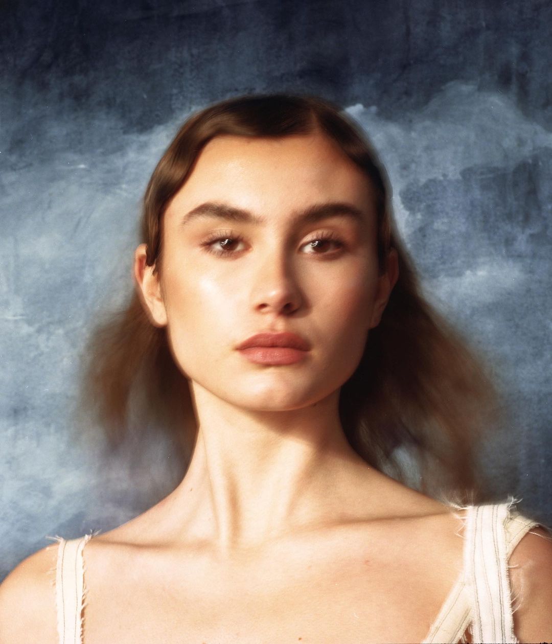 Close-up portrait of a fair-skinned young woman with brown hair and light makeup against a textured dark blue background in a studio setting.