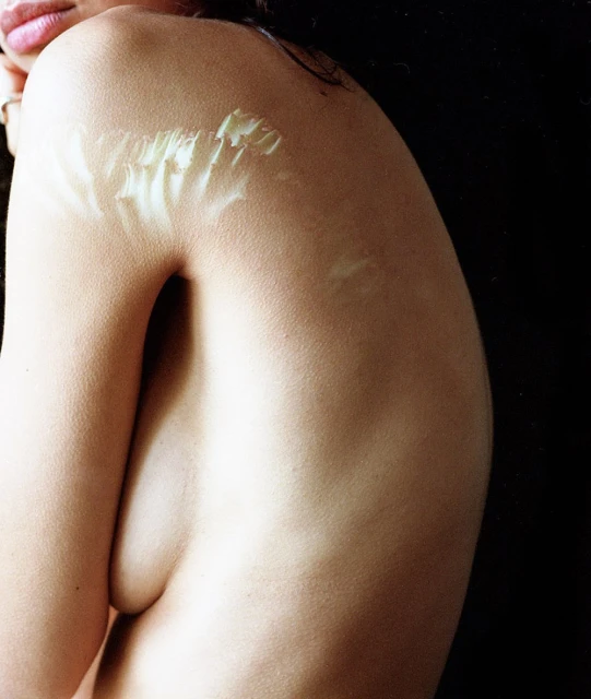 Close-up studio shot of a topless adult female with arm crossing chest. Fair skin is visible against a dark background.