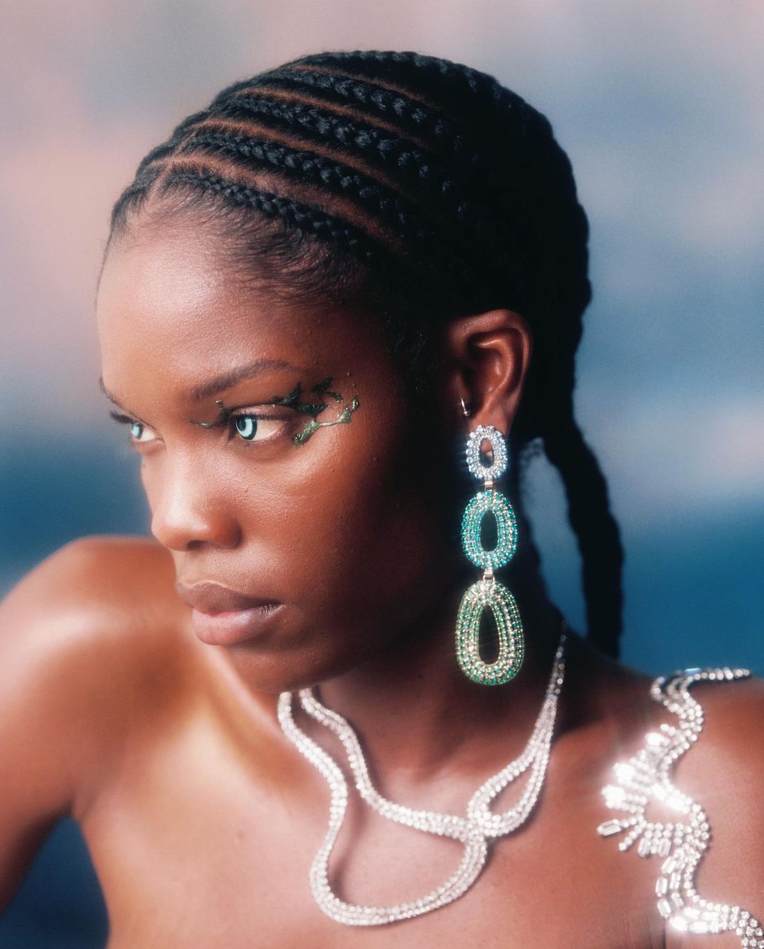 Close-up portrait of a woman with cornrow braids, wearing jeweled makeup, statement earrings, and a necklace against a blurred background.