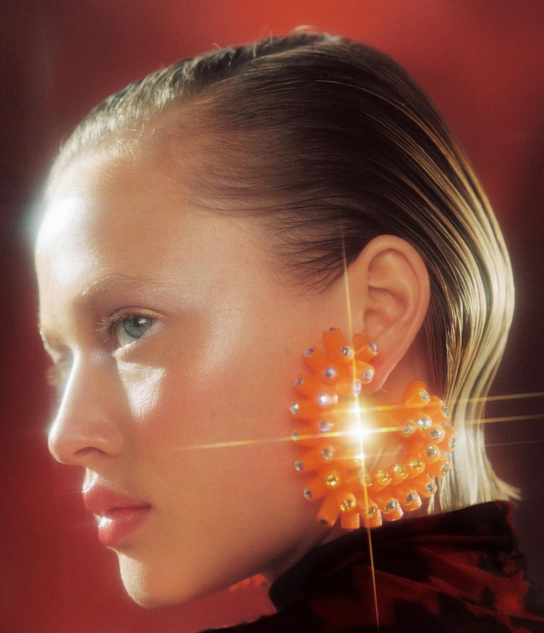 Close-up of a model with slicked-back hair wearing an orange heart-shaped earring with rhinestones against a warm reddish background.