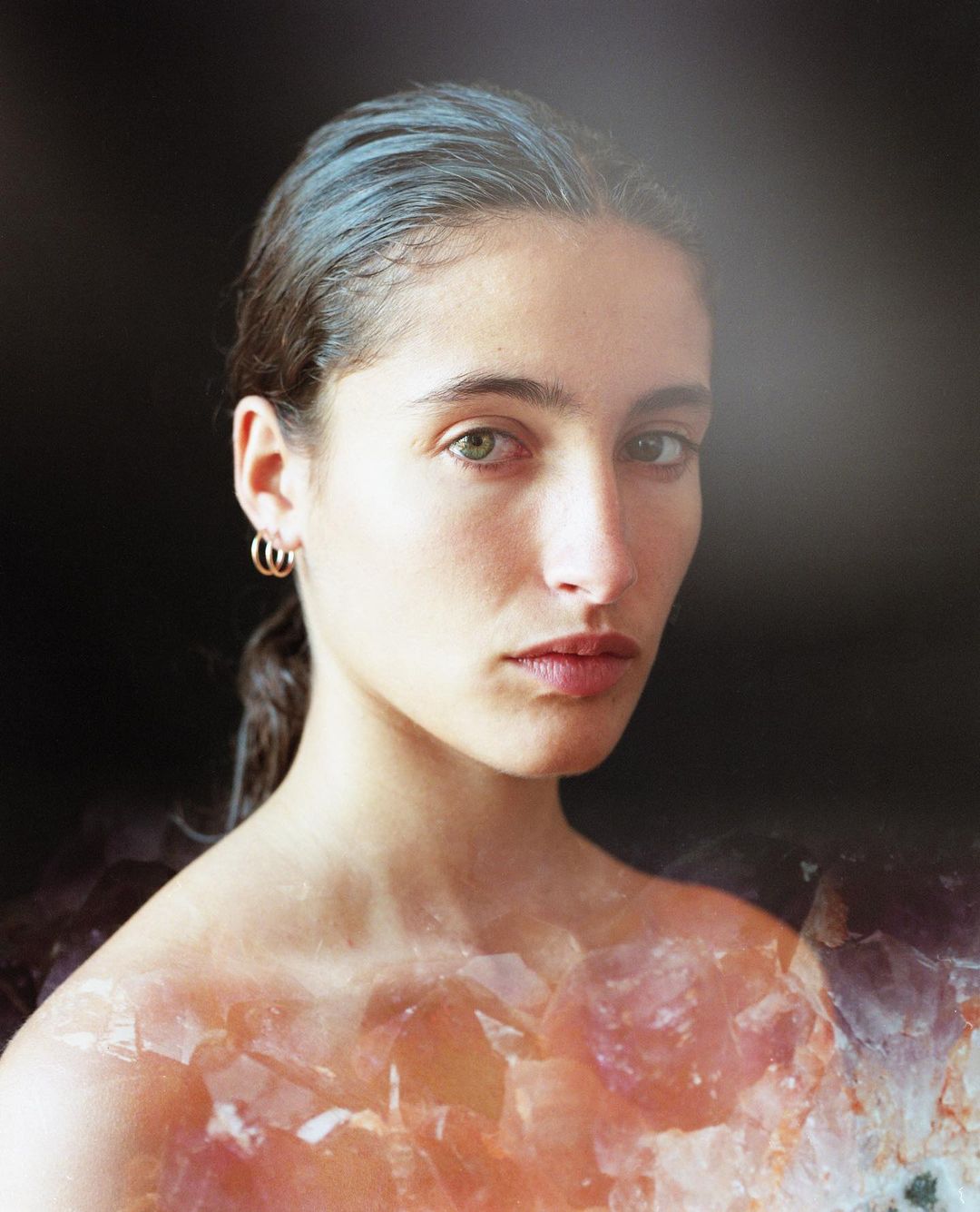 Close-up portrait of a woman with slicked-back dark hair and small gold hoop earrings layered over a crystalline amethyst cluster.