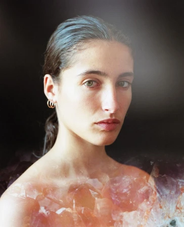 Close-up portrait of a woman with slicked-back dark hair and small gold hoop earrings layered over a crystalline amethyst cluster.