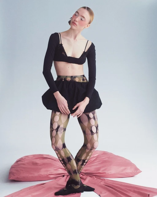 A fair-skinned woman poses in a black bra top, long-sleeved shirt, and ruffled skirt, accented by patterned tights and a pink bow.