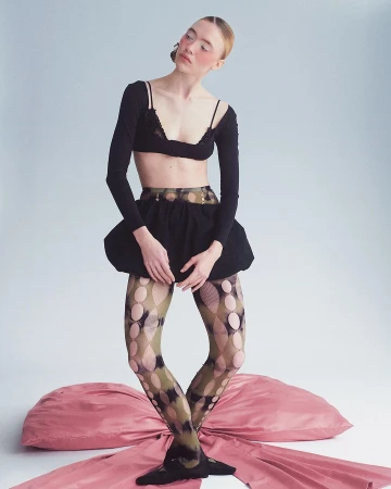 A fair-skinned woman poses in a black bra top, long-sleeved shirt, and ruffled skirt, accented by patterned tights and a pink bow.