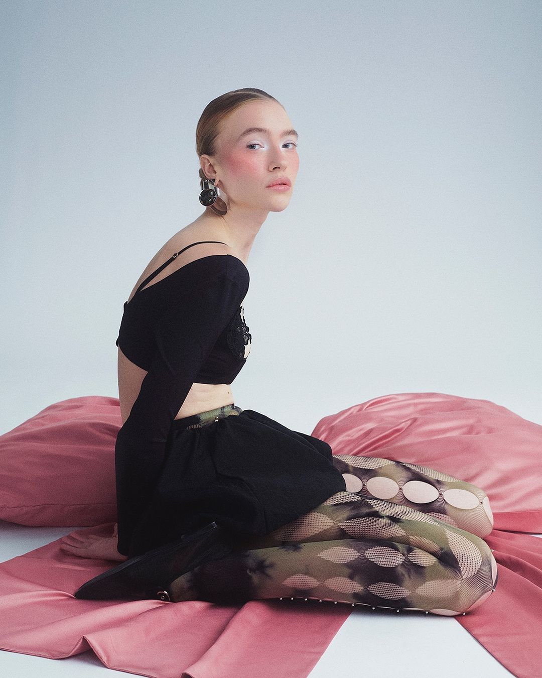 A young adult woman poses in a studio with black top and patterned tights, seated on rose-colored pillows against a neutral background.