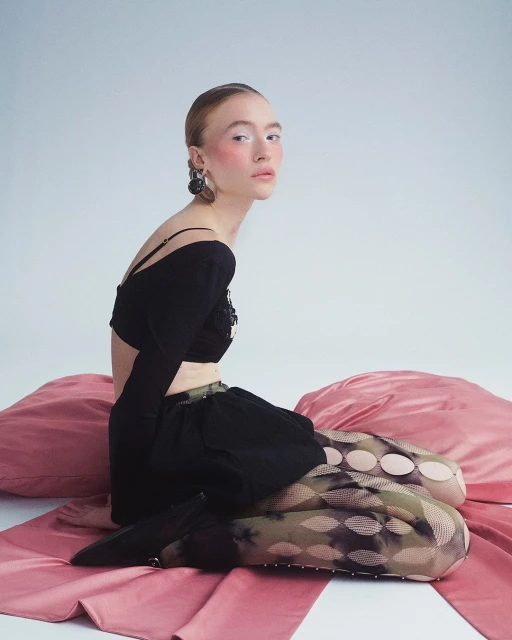 A young adult woman poses in a studio with black top and patterned tights, seated on rose-colored pillows against a neutral background.