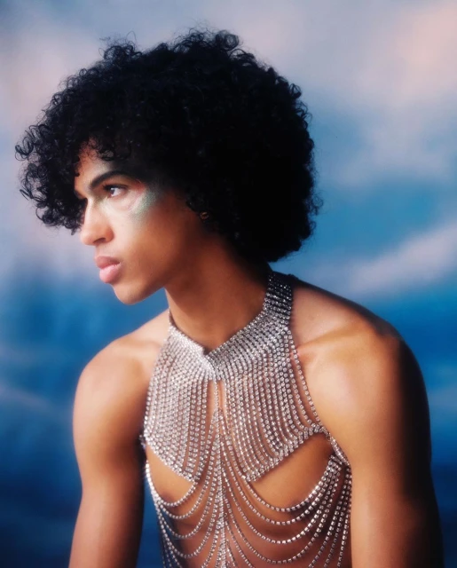 Portrait of a young adult male with curly hair wearing a rhinestone halter top against a blue and white cloud-like background.