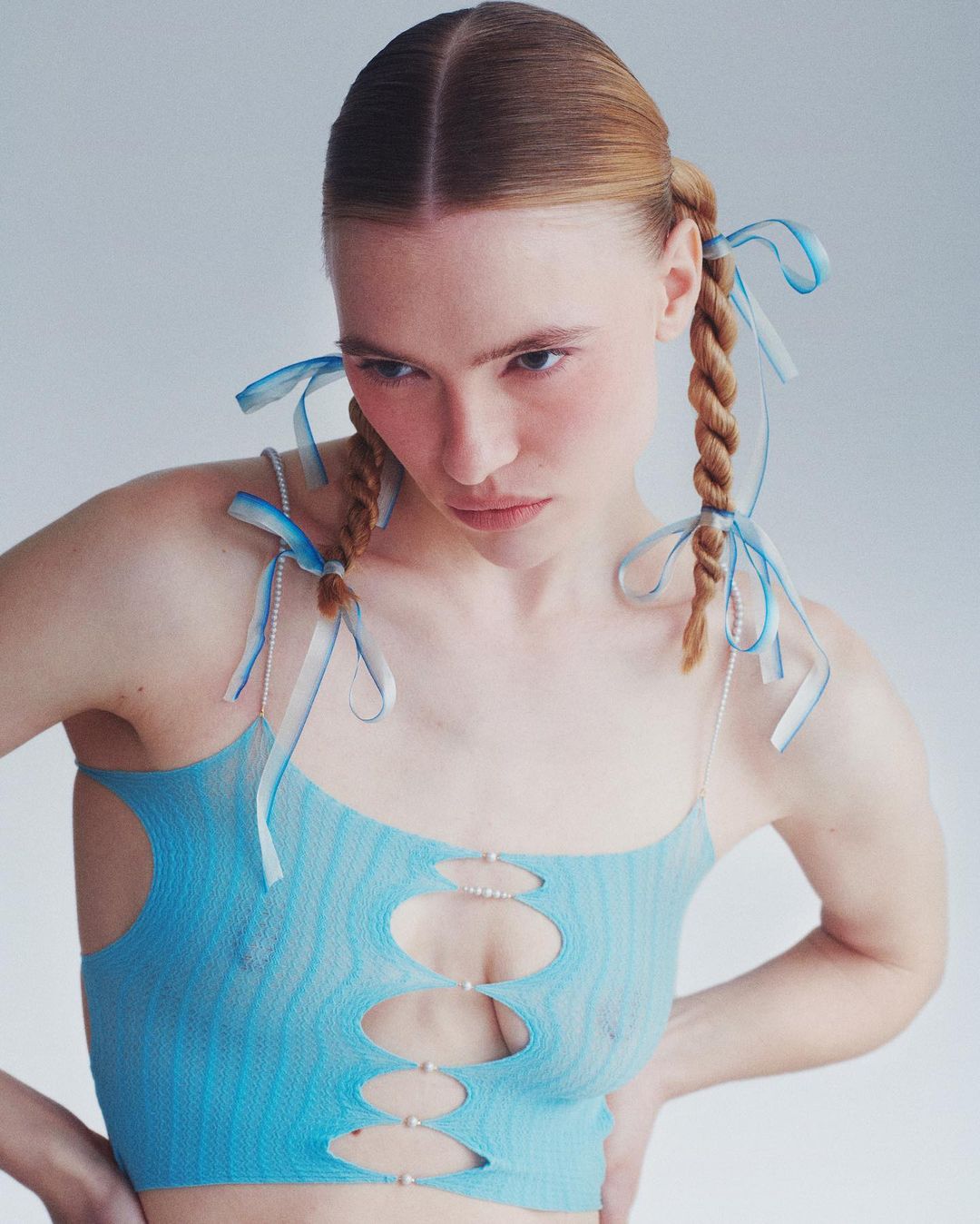 A young woman with braided hair and blue ribbons poses indoors in a light blue top with decorative pearls and cut-out details against a light grey background.