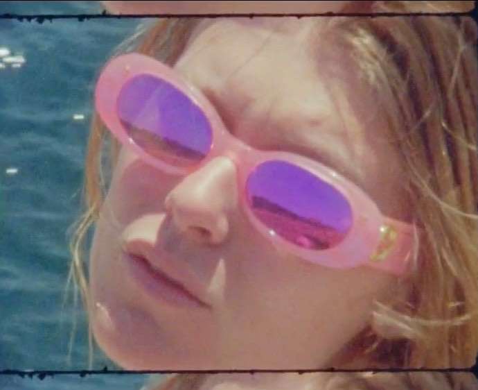 Close-up of a woman with light hair wearing pink-rimmed sunglasses that reflect the sky, positioned against a blurred blue background.