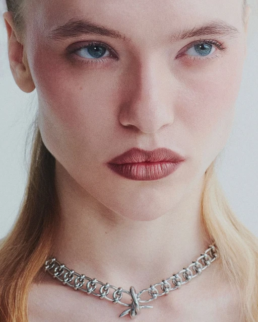 Close-up portrait of a fair-skinned woman with blue eyes and light brown hair wearing a silver chain necklace against a white background.