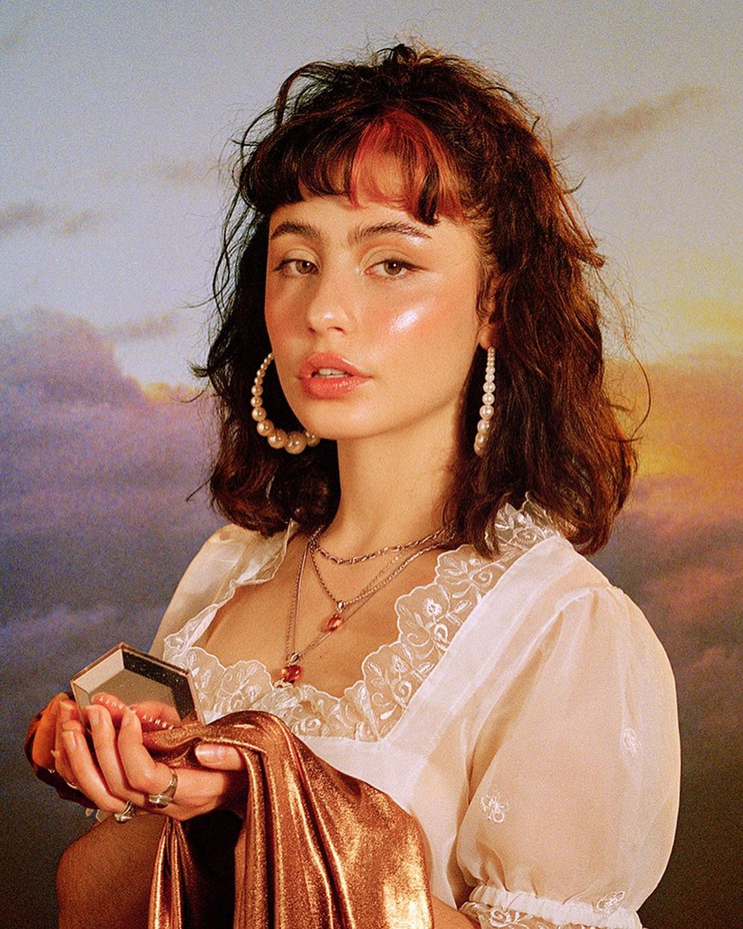 Brunette woman with bangs in a sheer white top holds gold fabric against a painted sky background, wearing layered necklaces and pearl earrings.